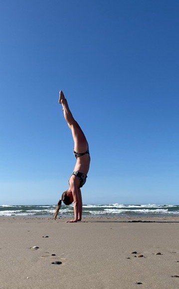 Eine Frau macht bei blauem Himmel einen Handstand am Strand, im Hintergrund das Meer