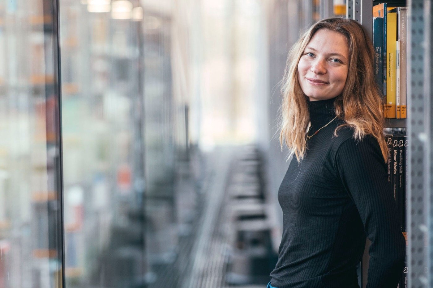 Stephi Bünemann lehnt lächelnd an einem Bücherregal in einer Bibliothek
