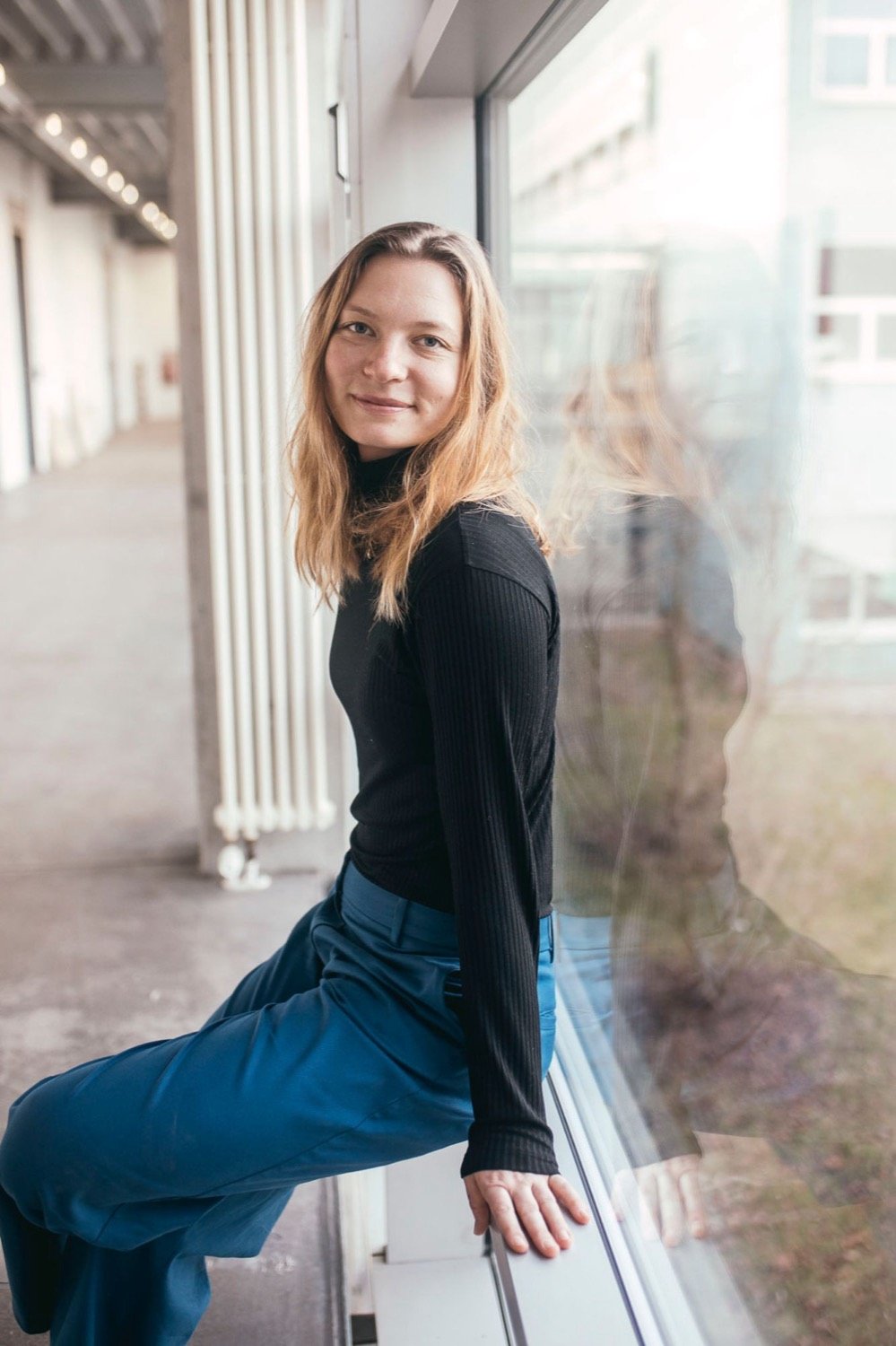 Stephi Bünemann sitzt in schwarzem Pullover und blauer Hose auf einer Fensterbank und schaut lächelnd über die Schulter