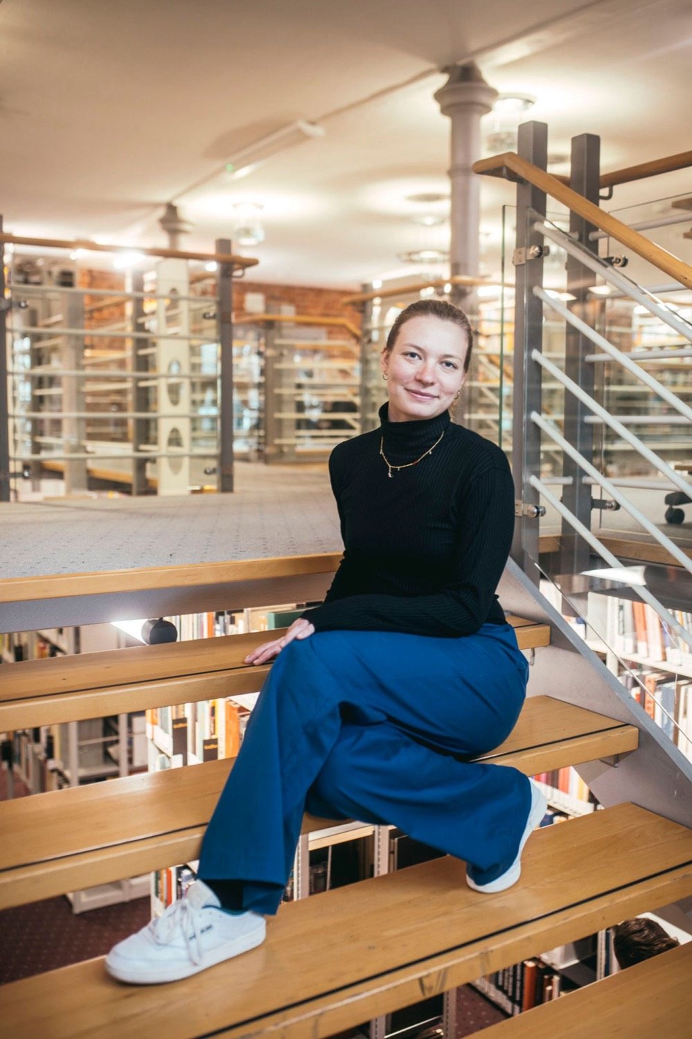Stephi Bünemann sitzt entspannt auf einer Holztreppe in einer Bibliothek und lächelt in die Kamera
